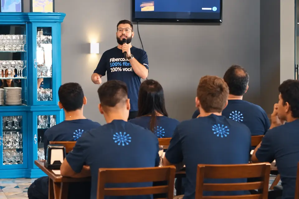 Mauro Fagotti, de pé e com microfone, fala para a equipe de um provedor sentada de costas, todos de camiseta azul igual.