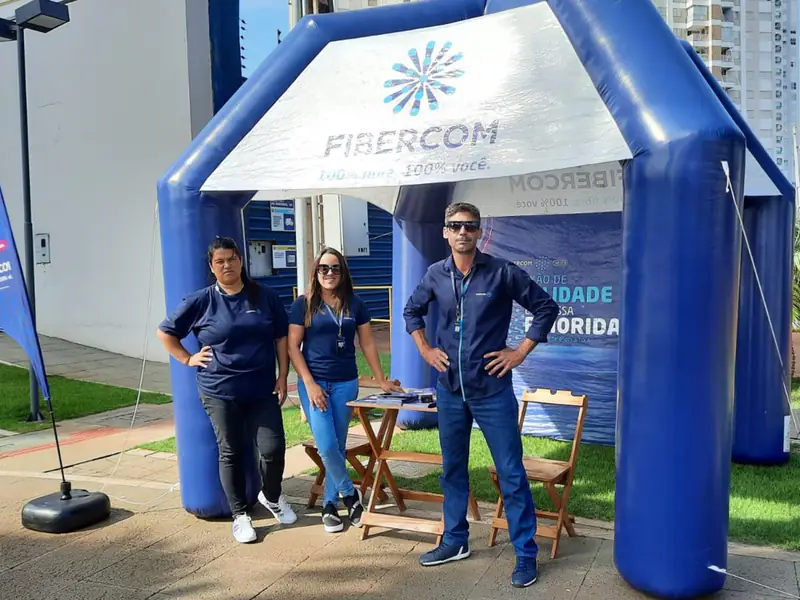 Tenda inflável da Fibercom num gramado, com três pessoas da equipe de vendas de uniforme azul.