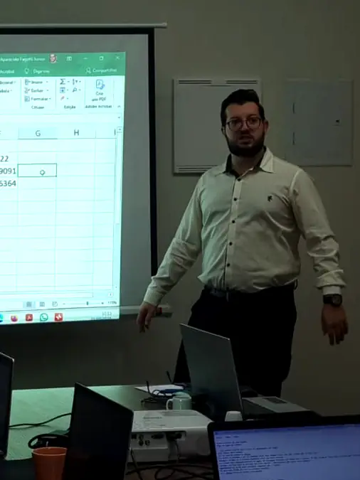 Mauro de pé ao lado de um telão com planilha de ativações e suporte, numa sala de reunião.