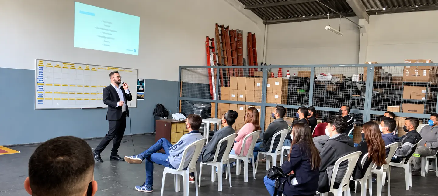 Mauro Fagotti, de terno e microfone, fala para a equipe de um provedor sentada num galpão. Na parede ao fundo, um quadro de metas.